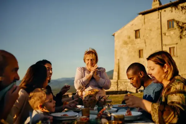 A group sharing a long table of food outdoors in the evening sun, an old stone farmhouse behind them